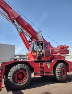 A large red mobile crane with oversized tires and an extended boom, situated outdoors against a clear blue sky. The machine has a cabin with visible controls and safety labels attached to various parts of its body.