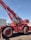 A large red mobile crane with oversized tires and an extended boom, situated outdoors against a clear blue sky. The machine has a cabin with visible controls and safety labels attached to various parts of its body.