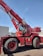 A large red mobile crane with oversized tires and an extended boom, situated outdoors against a clear blue sky. The machine has a cabin with visible controls and safety labels attached to various parts of its body.