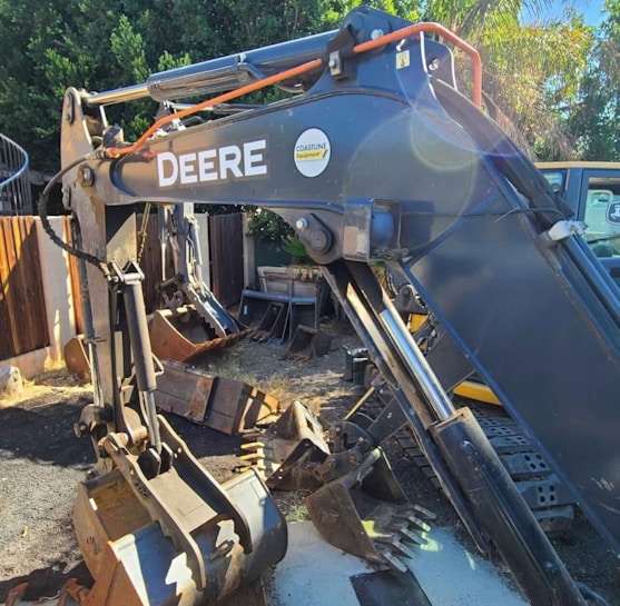 A close-up view of a construction excavator's arm and bucket is visible. The equipment is placed outdoors on a dirt-covered surface with some greenery in the background. The sunlight creates a lens flare on the machinery.