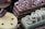 An assortment of elegantly crafted desserts is displayed, including a layered chocolate cake topped with candied nuts and dusted with powdered sugar, a yellow glazed mousse cake with chocolate garnish, and a light green pastry with decorative swirls on top.