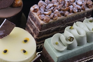 An assortment of elegantly crafted desserts is displayed, including a layered chocolate cake topped with candied nuts and dusted with powdered sugar, a yellow glazed mousse cake with chocolate garnish, and a light green pastry with decorative swirls on top.