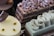 An assortment of elegantly crafted desserts is displayed, including a layered chocolate cake topped with candied nuts and dusted with powdered sugar, a yellow glazed mousse cake with chocolate garnish, and a light green pastry with decorative swirls on top.