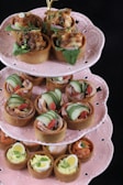 An elegant three-tiered stand holds an assortment of savory tarts, each on a delicate pink ceramic dish. The top tier displays tarts with a crispy brown crust, topped with green leaves, herbs, and a savory filling. The middle tier features tarts garnished with sliced cucumber and red berries, suggesting fresh and colorful ingredients. The bottom tier includes tarts with hard-boiled quail eggs and a creamy filling, indicating a rich flavor profile.
