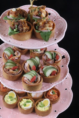 An elegant three-tiered stand holds an assortment of savory tarts, each on a delicate pink ceramic dish. The top tier displays tarts with a crispy brown crust, topped with green leaves, herbs, and a savory filling. The middle tier features tarts garnished with sliced cucumber and red berries, suggesting fresh and colorful ingredients. The bottom tier includes tarts with hard-boiled quail eggs and a creamy filling, indicating a rich flavor profile.