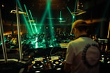 A DJ stands behind mixing equipment, performing in a dimly lit club filled with a large, energetic crowd. Colored lights and beams illuminate the scene, creating a lively atmosphere.