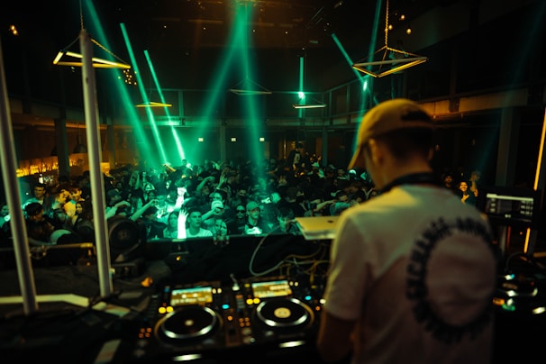A DJ stands behind mixing equipment, performing in a dimly lit club filled with a large, energetic crowd. Colored lights and beams illuminate the scene, creating a lively atmosphere.