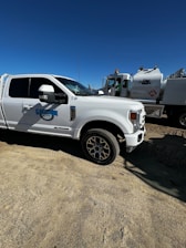 A white utility truck with branding on the side is parked on a sandy surface. In the background, another truck with a large tank and construction company logo is visible. The sky is clear and blue.