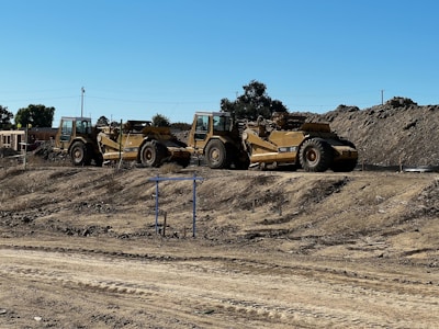 Two large construction vehicles, specifically scrapers, are parked on a dirt construction site. The ground is dry and uneven, with visible tread marks. Piles of earth are seen in the background, along with trees and a clear blue sky.