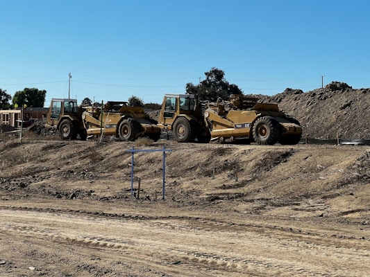 Two large construction vehicles, specifically scrapers, are parked on a dirt construction site. The ground is dry and uneven, with visible tread marks. Piles of earth are seen in the background, along with trees and a clear blue sky.