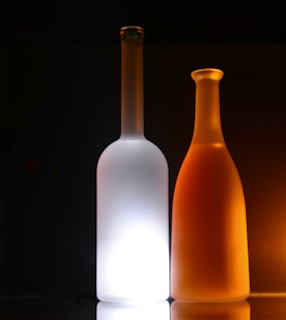 Two glass bottles with a minimalist aesthetic, one frosted white and the other a warm amber hue. They are positioned side by side against a dark background, with subtle reflections on the surface beneath them.