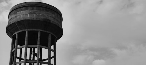 A large industrial water tower stands with its cylindrical tank mounted on top of a series of vertical supports against a partly cloudy sky. The structure is made of concrete and has a robust, imposing presence.
