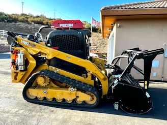 A yellow tracked skid steer loader with a mulching attachment, parked beside a building. There is an American flag in the background and a fire extinguisher mounted on the side of the machine.