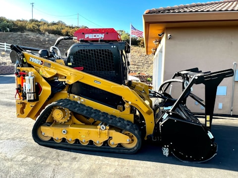 A yellow tracked skid steer loader with a mulching attachment, parked beside a building. There is an American flag in the background and a fire extinguisher mounted on the side of the machine.