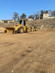 A construction site with large yellow machinery, including a bulldozer and a road roller, situated on a dirt ground. The background features a hillside with residential buildings and sparse trees under a clear blue sky.