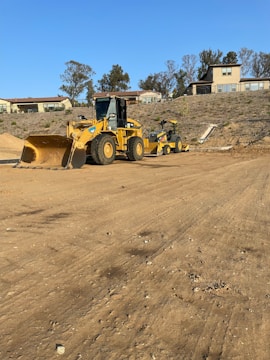 A construction site with large yellow machinery, including a bulldozer and a road roller, situated on a dirt ground. The background features a hillside with residential buildings and sparse trees under a clear blue sky.