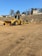 A construction site with large yellow machinery, including a bulldozer and a road roller, situated on a dirt ground. The background features a hillside with residential buildings and sparse trees under a clear blue sky.