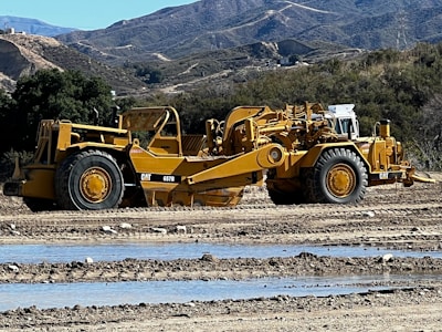 A large yellow construction vehicle is parked on a dirt road, with hilly terrain and trees in the background. The vehicle is a Caterpillar model with large tires and a long articulated arm.