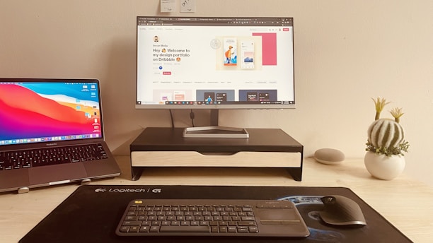 A computer setup featuring a MacBook on the left, and a large monitor on a stand in the center displaying a design portfolio. Below the monitor, a Logitech keyboard and mouse are placed on a black desk mat. A small cactus plant in a white pot and a gray Google Nest Mini are on the right side on a wooden desk.