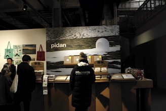 A group of people stands in front of a counter displaying various products. The background features a large image of a snowy landscape and the word 'pidan' is prominently displayed. The area appears to be indoors, likely a vendor stall or exhibition stand. The counter has boxes and products neatly arranged, with posters and bags hanging on the side wall. One person is wearing a black jacket with the word 'HERITAGE' on the back.