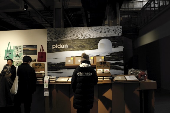 A group of people stands in front of a counter displaying various products. The background features a large image of a snowy landscape and the word 'pidan' is prominently displayed. The area appears to be indoors, likely a vendor stall or exhibition stand. The counter has boxes and products neatly arranged, with posters and bags hanging on the side wall. One person is wearing a black jacket with the word 'HERITAGE' on the back.