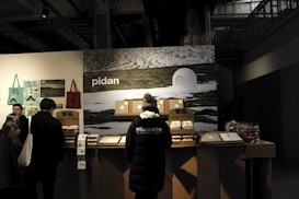 A group of people stands in front of a counter displaying various products. The background features a large image of a snowy landscape and the word 'pidan' is prominently displayed. The area appears to be indoors, likely a vendor stall or exhibition stand. The counter has boxes and products neatly arranged, with posters and bags hanging on the side wall. One person is wearing a black jacket with the word 'HERITAGE' on the back.