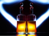 Two elegant glass bottles with spherical stoppers are placed side by side, containing a warm amber liquid. They are set against a dramatic black background, with white and blue light illuminating and reflecting off the glass, creating a striking symmetrical effect.