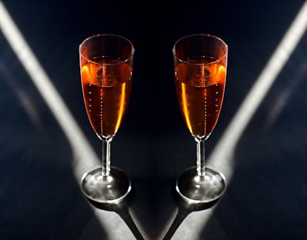 Two champagne glasses with an amber liquid are placed symmetrically against a dark background, illuminated by converging light beams. The glasses cast long shadows, creating a dramatic and elegant atmosphere.