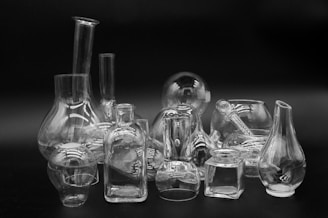 A collection of transparent glassware consisting of various shapes and sizes, including flasks, bottles, and jars, arranged on a flat surface against a dark background. The reflective surfaces of the glass create intricate patterns of light and shadow.