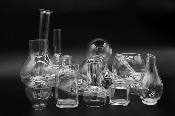A collection of transparent glassware consisting of various shapes and sizes, including flasks, bottles, and jars, arranged on a flat surface against a dark background. The reflective surfaces of the glass create intricate patterns of light and shadow.