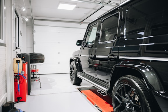 A sleek black SUV is parked inside a well-lit clean garage. To the left, there are multiple tools, a power outlet, and a set of spare tires. The garage door is closed, and the floor is spotless.