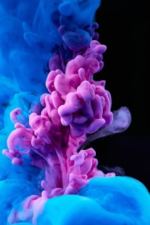Vibrant plumes of pink and blue ink swirl and mix together in water, creating an abstract and mesmerizing pattern against a dark background.