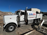 A large white industrial truck with "Calex Engineering Co." branding on the side is parked in an outdoor industrial setting. The truck has multiple axles and shiny metal tanks, indicating it may be used for specialized construction or engineering tasks. Additional buildings and equipment are visible in the background under a clear blue sky.