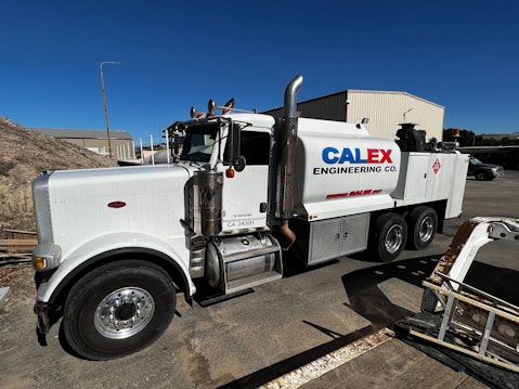 A large white industrial truck with "Calex Engineering Co." branding on the side is parked in an outdoor industrial setting. The truck has multiple axles and shiny metal tanks, indicating it may be used for specialized construction or engineering tasks. Additional buildings and equipment are visible in the background under a clear blue sky.