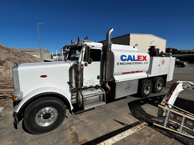 A large white industrial truck with "Calex Engineering Co." branding on the side is parked in an outdoor industrial setting. The truck has multiple axles and shiny metal tanks, indicating it may be used for specialized construction or engineering tasks. Additional buildings and equipment are visible in the background under a clear blue sky.