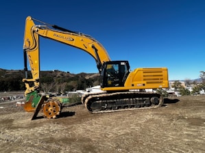 A large yellow excavator is positioned on a construction site with a clear blue sky in the background. The machine has a robust tracked base and a large articulated arm with a hydraulic attachment. Surrounding the site are rolling hills and some industrial buildings.