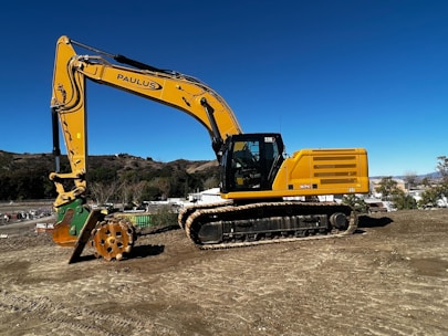 A large yellow excavator is positioned on a construction site with a clear blue sky in the background. The machine has a robust tracked base and a large articulated arm with a hydraulic attachment. Surrounding the site are rolling hills and some industrial buildings.