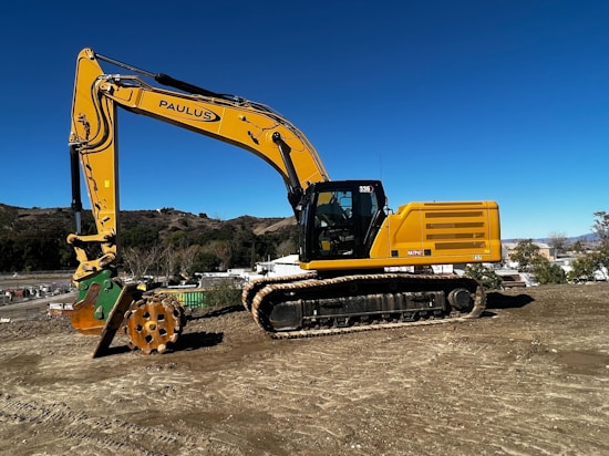 A large yellow excavator is positioned on a construction site with a clear blue sky in the background. The machine has a robust tracked base and a large articulated arm with a hydraulic attachment. Surrounding the site are rolling hills and some industrial buildings.