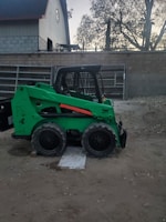 A skid-steer loader parked on a sandy path with tropical foliage in the background.