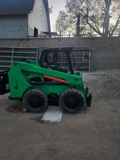 A green skid steer loader with black wheels is parked on a dirt surface. The vehicle has an enclosed cab and is positioned near a brick wall with a large building and trees visible in the background. The setting appears to be a semi-rural or construction area, with leaves scattered across the ground.