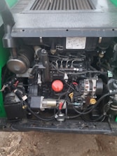 A close-up view of an engine compartment with various mechanical components and wiring. The engine features metallic parts, a red circular cap, and a yellow dipstick. Surrounding parts include hoses, connectors, and a black battery. The setup is enclosed in a dark-colored frame with bolts and labels visible.