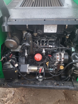 A close-up view of an engine compartment with various mechanical components and wiring. The engine features metallic parts, a red circular cap, and a yellow dipstick. Surrounding parts include hoses, connectors, and a black battery. The setup is enclosed in a dark-colored frame with bolts and labels visible.