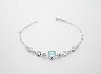 A delicate silver bracelet featuring a central heart-shaped blue gemstone flanked by two smaller round white stones on each side, all linked with a fine chain.