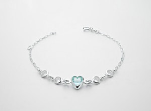 A delicate silver bracelet featuring a central heart-shaped blue gemstone flanked by two smaller round white stones on each side, all linked with a fine chain.