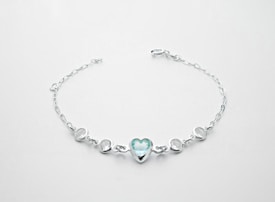 A delicate silver bracelet featuring a central heart-shaped blue gemstone flanked by two smaller round white stones on each side, all linked with a fine chain.