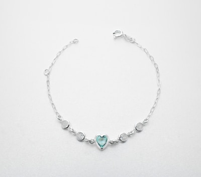 Delicate silver bracelet with a heart-shaped blue gemstone and four round clear gemstones evenly spaced along the chain. It has a simple chain link design and a clasp for securing it.