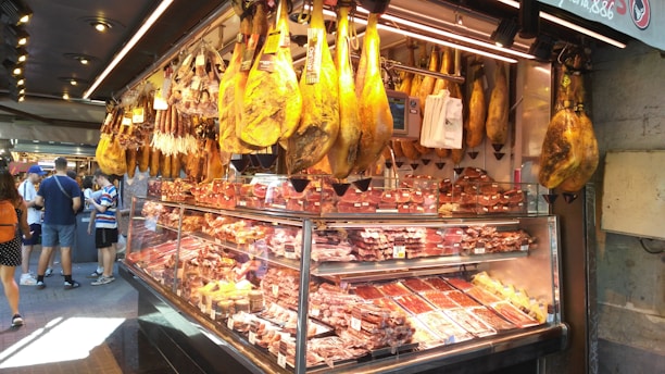 A market stall displaying a wide variety of cured meats, including large hams hanging from the ceiling and multiple cuts of meat within the glass counter. Several customers are present, observing the selection.