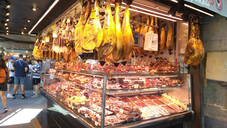 A market stall displaying a wide variety of cured meats, including large hams hanging from the ceiling and multiple cuts of meat within the glass counter. Several customers are present, observing the selection.