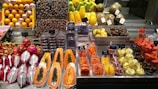 A vibrant display of fruits at a market, featuring an array of colorful produce including dragon fruits with spoons, papayas, melons, dates, and assorted fruit slices neatly packaged. Price tags are visible, indicating a commercial setting with various types of fresh and possibly exotic fruits.