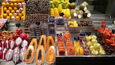 A vibrant display of fruits at a market, featuring an array of colorful produce including dragon fruits with spoons, papayas, melons, dates, and assorted fruit slices neatly packaged. Price tags are visible, indicating a commercial setting with various types of fresh and possibly exotic fruits.